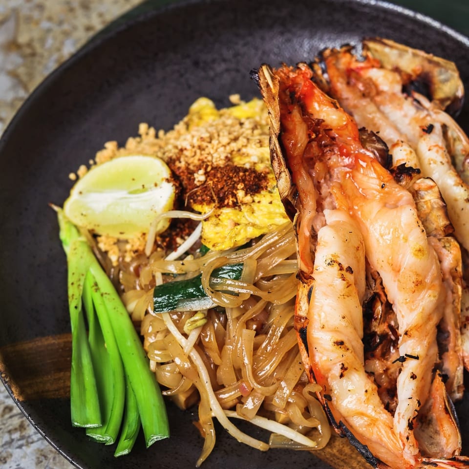 Chao Krung Thai's Pad See Ew with grilled prawns, lime, and green onions on a dark plate.